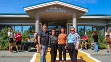 Crosswalk - Board Chair group photo 1 man and 3 female leadership team members pose in front of elementary school and on new orange crosswalk