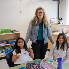 Female Educational Assistant stands with two female elementary students who are eating lunch