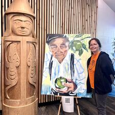 A women stands next to the painting of her father and a carving the represents her brother